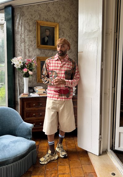 Camisa de cuadros en rojo y beige con un parche estampado, pantalones cortos beige y zapatillas beige. Fondo: jarrón con flores, cajón de madera y una obra de arte.