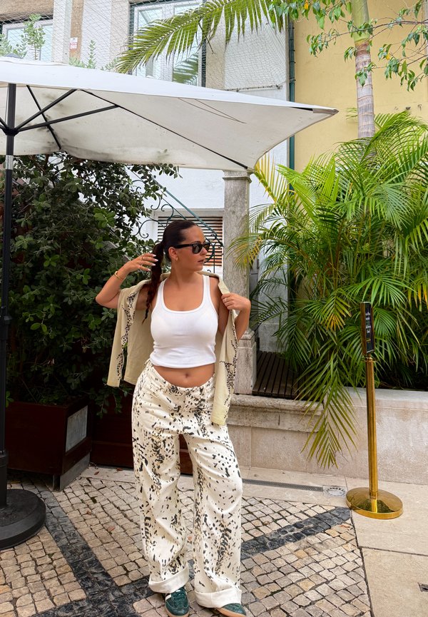 Femme aux cheveux tressés portant des lunettes de soleil, un débardeur blanc, un pantalon à motifs et une veste, posant sur une terrasse carrelée avec un parasol et des plantes vertes à proximité.