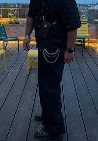 Chemise noire à manches courtes avec des détails à clous, pantalon noir avec des accents en chaîne et chaussures noires. Se tenant sur des planches en bois foncé.