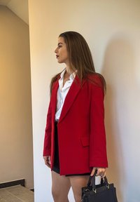 Rød skreddersydd blazer med skarpe revers, sidelommer og strukturert passform, kombinert med en hvit skjorte og en svart håndveske.