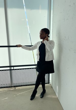 Una mujer con un cárdigan blanco sobre un vestido negro, medias negras y zapatos negros, apoyada en una barandilla en un espacio interior brillante.