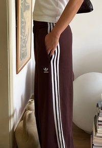 Bordové tepláky Adidas s tromi bielymi pruhmi pozdĺž strán, obsahujúce malé logo na ľavom stehne a bočnú kapsu.