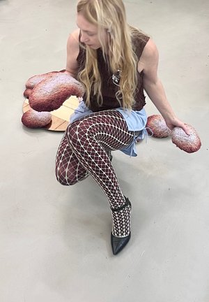 Hasta las rodillas en una falda azul claro, llevando medias de red color marrón y tacones negros de punta, sosteniendo objetos texturizados con formas orgánicas.