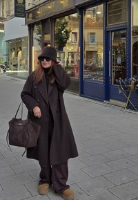 Long manteau marron foncé sur une tenue noire, grand chapeau marron surdimensionné, lunettes de soleil et sac marron texturé ; debout sur un pavé en pierre.
