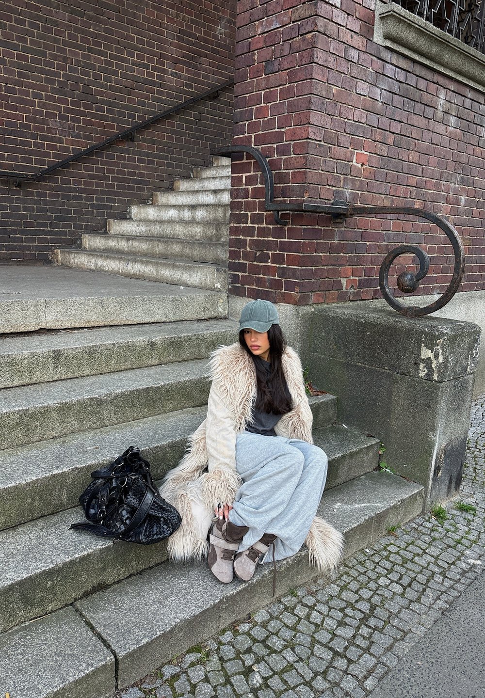 Jeune femme aux longs cheveux noirs, vêtue d'un manteau en fourrure et d'une casquette grise, assise sur des marches en pierre à côté d'un grand sac noir près d'un bâtiment en briques.