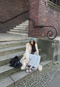 Jeune femme aux longs cheveux noirs, vêtue d'un manteau en fourrure et d'une casquette grise, assise sur des marches en pierre à côté d'un grand sac noir près d'un bâtiment en briques.
