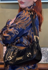 Femme aux cheveux roux portant un chemisier bleu et marron à motifs, portant un sac à bandoulière en cuir noir Versace Couture avec des boucles dorées.