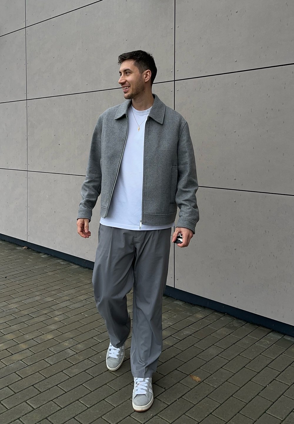 Grey zip-up jacket with a textured finish, white t-shirt underneath, loose grey trousers, and grey trainers on a paved surface.