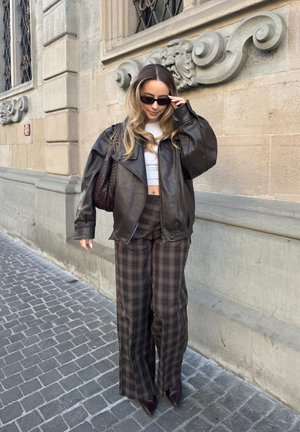 Mujer con chaqueta de cuero oscura, top corto blanco, pantalones anchos de cuadros, gafas de sol y zapatos puntiagudos, de pie en una calle empedrada junto a un edificio de piedra.
