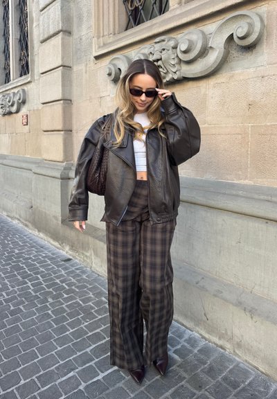 Mujer con chaqueta de cuero oscura, top corto blanco, pantalones anchos de cuadros, gafas de sol y zapatos puntiagudos, de pie en una calle empedrada junto a un edificio de piedra.
