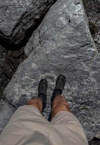 Isik, kes kannab beežikaid lühikesi pükse ja musti matkajalatsid, seisab välitingimustes suurel hallil kivisel pinnal.