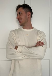 T-shirt à manches longues côtelées de couleur crème avec un col rond, arborant un petit détail de texte brodé en or sur la poitrine.