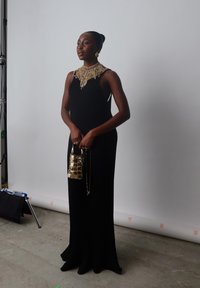 Czarna wieczorowa suknia z prążkowanej tkaniny z efektownym złotym naszyjnikiem z koralików. Metaliczna złota torebka z łańcuszkiem. Gładkie tło.