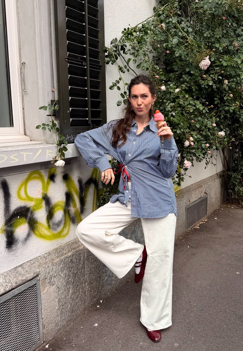 Femme en chemise rayée et pantalon blanc tenant un cornet de glace rose, posant avec un pied levé près d’un mur couvert de graffitis et de buissons de roses.