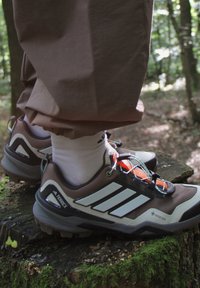 Brūni pārgājienu apavi ar baltiem akcentiem, GORE-TEX zīmolu un oranžiem šņores. Teksturotas gumijas zoles labai saķerei, valkāti ar baltām zeķēm.