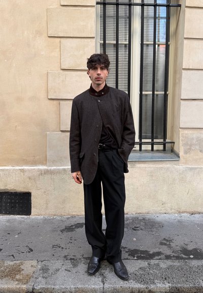Joven con cabello rizado y oscuro, vestido con un blazer gris, camisa negra, pantalones negros y zapatos negros, parado en una acera junto a una pared beige.