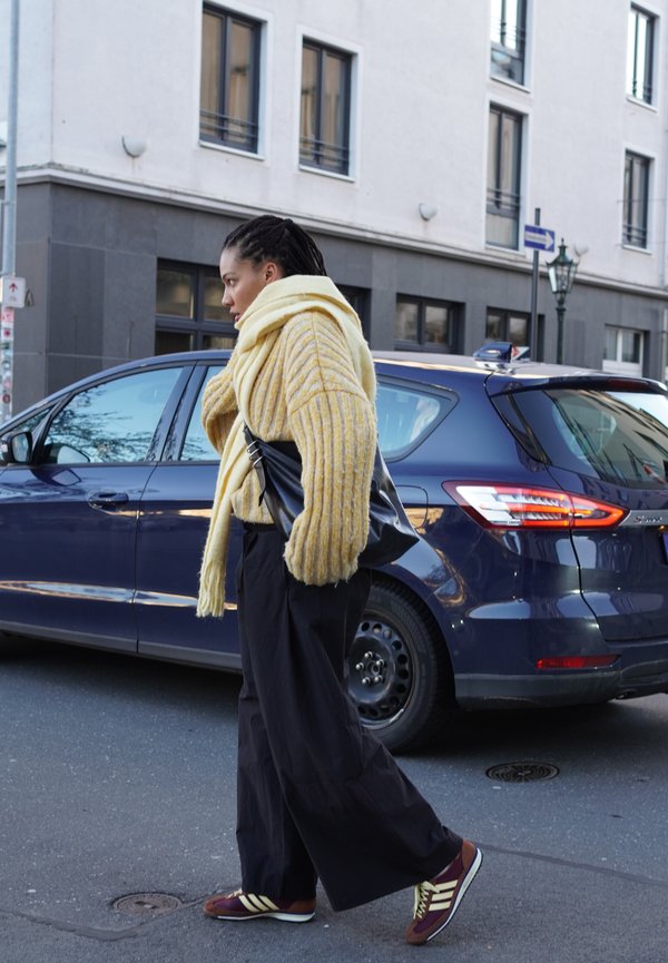 Żółty sweter w paski, oversize, o prążkowanej fakturze; czarne szerokie spodnie; bordowe trampki z żółtymi akcentami; idący obok niebieskiego samochodu.