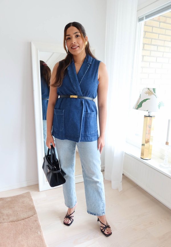 Vrouw draagt een mouwloos blauw vest, lichte jeans en zwarte hakken, houdt een zwarte handtas vast, staat binnen bij een spiegel en raam met lamp.