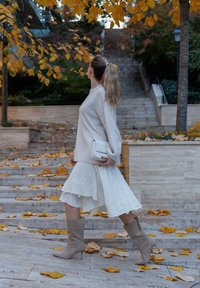 Svijetlosmeđi pulover, bijela slojevita suknja s cvjetnim vezom, bež čizmice do koljena i mala bijela torbica. Lišće raspršeno po stepenicama.