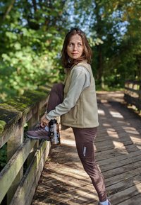 Beige Fleece-Weste über einem hellen Langarmoberteil, braune enge Hose und graue Wanderschuhe. Hält eine Wasserflasche, während er/sie sich auf einer Holzbrücke dehnt.
