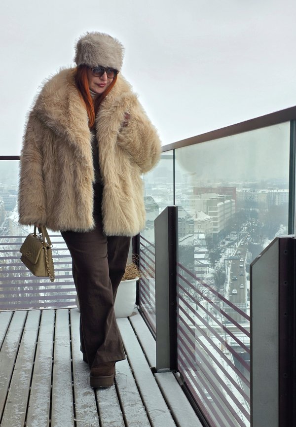 Donna con cappotto di pelliccia beige e cappello cammina su un balcone innevato che si affaccia su una strada della città attraverso una ringhiera di vetro.