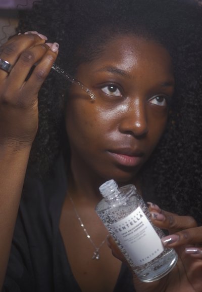 Mujer con cabello rizado aplicando suero facial con un cuentagotas cerca de su ojo, sosteniendo una botella transparente etiquetada con líquido en su interior.