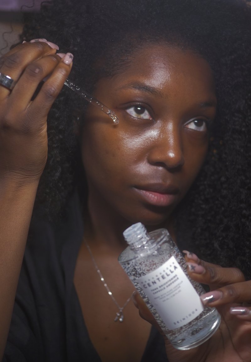 Femme aux cheveux bouclés appliquant un sérum facial à l'aide d'un compte-gouttes près de son œil, tenant un flacon transparent étiqueté contenant du liquide.