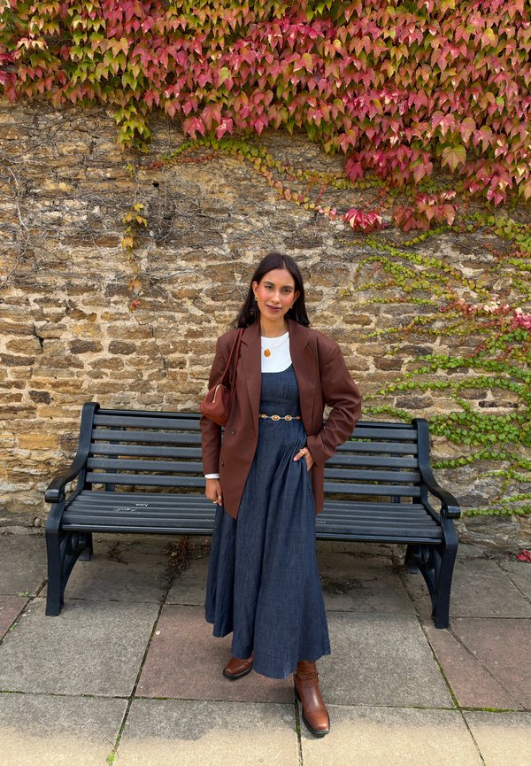 Femme portant un blazer marron et une robe bleu foncé se tient devant un mur de pierre avec du lierre rouge et vert, à côté d'un banc noir en plein air.