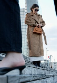 Cappotto lungo in camoscio marrone sopra una gonna bianca strutturata, abbinato a stivali neri alti al ginocchio. Borsa marrone minimalista e occhiali da sole scuri.