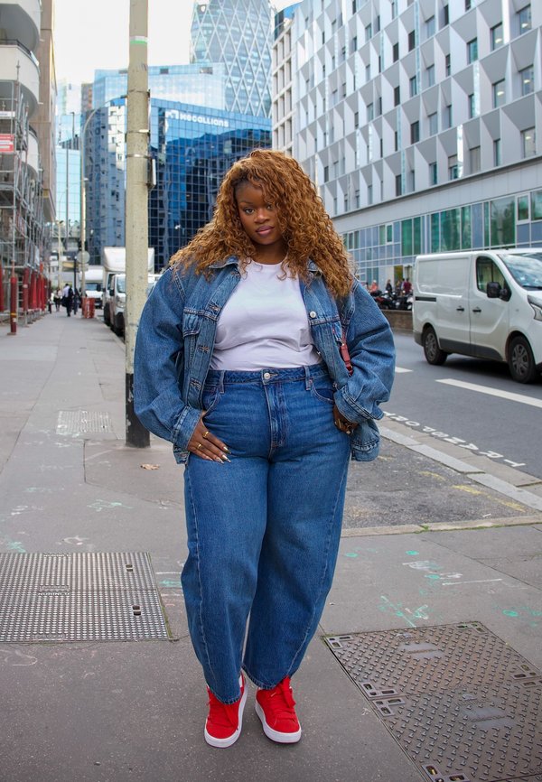 Une femme en veste en jean, en jeans et en baskets rouges se tient sur un trottoir en ville, parmi des immeubles de bureaux modernes et des camionnettes garées.