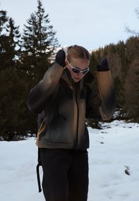 Personne portant des lunettes de soleil, une veste d'hiver et des moufles ajustant sa capuche en se tenant sur la neige avec des arbres à feuilles persistantes en arrière-plan.