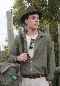 Jeune homme portant un chapeau vert et une chemise verte, un t-shirt blanc, un pantalon beige, portant un sac à dos vert avec une bouteille d'eau et une canette, debout près d'une clôture en bois avec de la verdure.