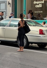 Žena v čiernom oblečení a sandáloch stojí pri zaparkovanom béžovom aute pred kaviarňou doogood, s ľuďmi vo vnútri a na chodníku v blízkosti.
