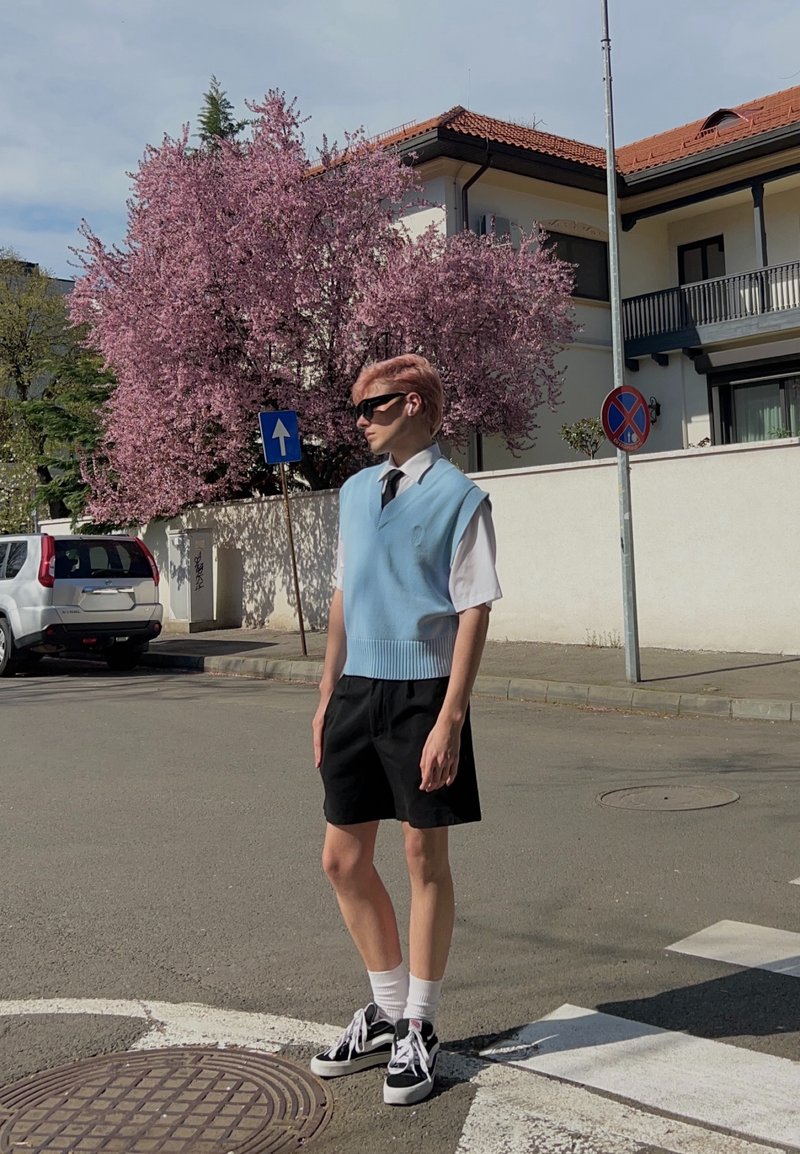 Jeune personne portant des lunettes de soleil, un gilet bleu clair, une chemise blanche, un short noir et des baskets, debout au coin de la rue près d'un arbre rose en fleurs et des panneaux de signalisation.