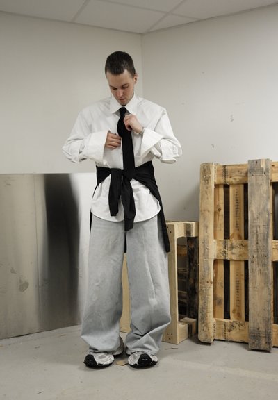 Camisa blanca de botones con grandes puños, corbata negra y pantalones grises sueltos. Cinturón negro atado en la cintura. Fondo neutro con palets de madera.