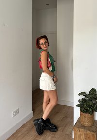 Πράσινο χωρίς μανίκια τοπ, λευκή μίνι φούστα με τσέπες, μαύρα χοντρά αθλητικά παπούτσια, κόκκινη τσάντα ώμου με κρόσια, oversized γυαλιά ηλίου, εσωτερικός χώρος.