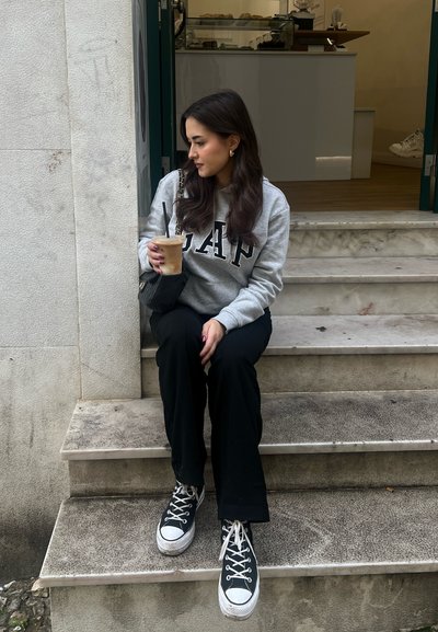 Sudadera gris con el logo "GAP", pantalones de pierna ancha negros, bolso negro y zapatillas en blanco y negro, sentada en escaleras de piedra con café helado.