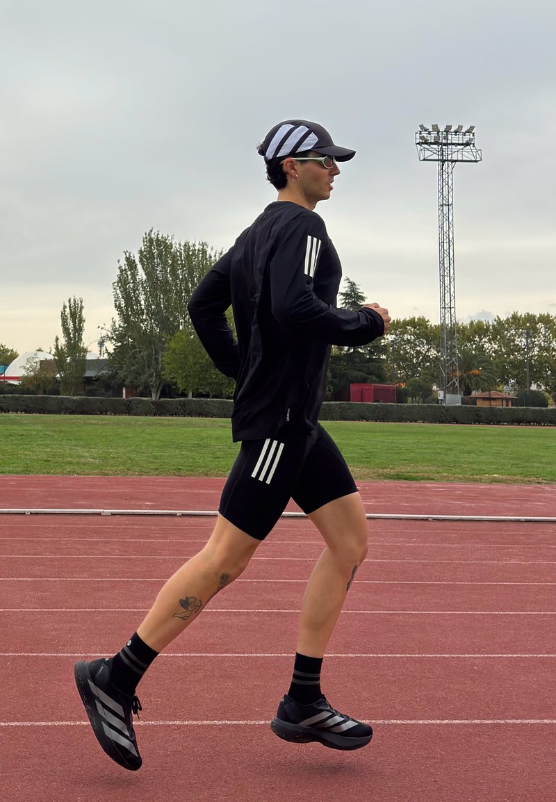 Un coureur vêtu de vêtements de sport noirs avec des rayures blanches, une casquette noire et des lunettes de soleil, joggant sur une piste extérieure rouge avec des arbres et un projecteur en arrière-plan.