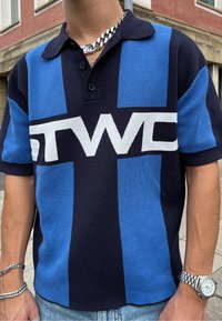 Zils un tumši zils polo krekls ar lielu baltu "STWC" logo, īsām piedurknēm, strukturētu trikotāžas audumu un klasisku apkakli.