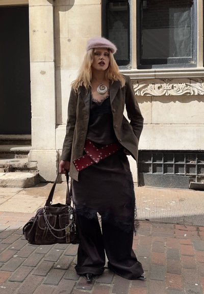 Mujer con boina rosa, chaqueta marrón, vestido negro de encaje sobre pantalones anchos, cinturón rojo con tachuelas, sujetando un gran bolso marrón en la acera de la ciudad.