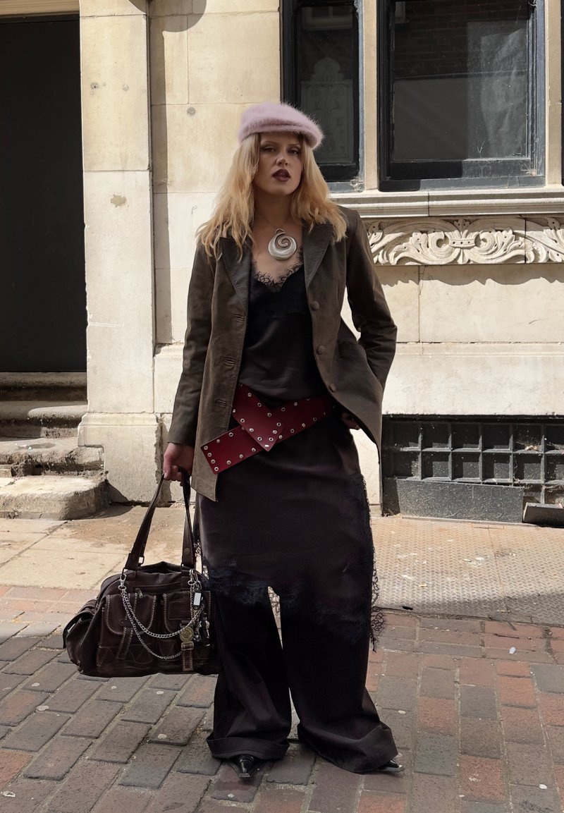 Femme portant un béret rose, une veste marron, une robe en dentelle noire sur un pantalon large, une ceinture rouge cloutée, tenant un grand sac à main marron sur un trottoir en ville.