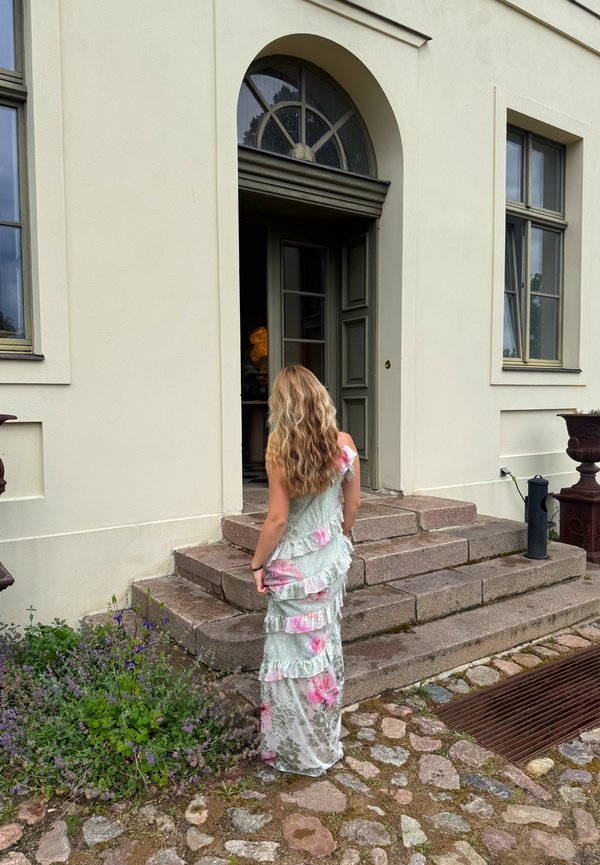 Vrouw met lang golvend haar in een pastelkleurige bloemenjurk staat op stenen trappen en kijkt naar een open deur van een lichtgekleurde gebouw met een boogramen.