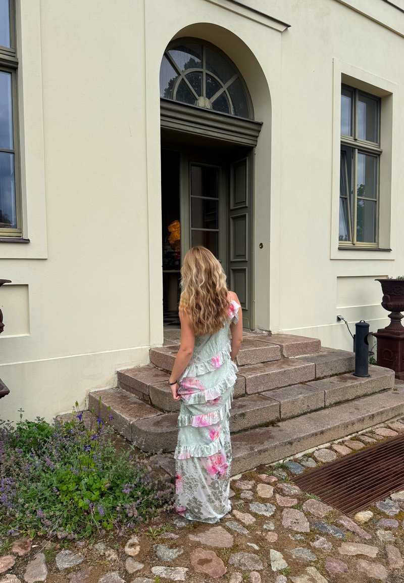 Femme aux longs cheveux ondulés portant une robe florale pastel, debout sur des marches en pierre face à une porte ouverte d'un bâtiment clair avec une fenêtre en arc.