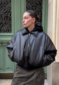 Νεαρός με τα μαλλιά χτενισμένα προς τα πίσω, τατουάζ στον λαιμό, φορώντας υπερμεγέθη μαύρο δερμάτινο μπουφάν και χακί παντελόνι, στέκεται μπροστά σε μια πράσινη διακοσμητική πόρτα.