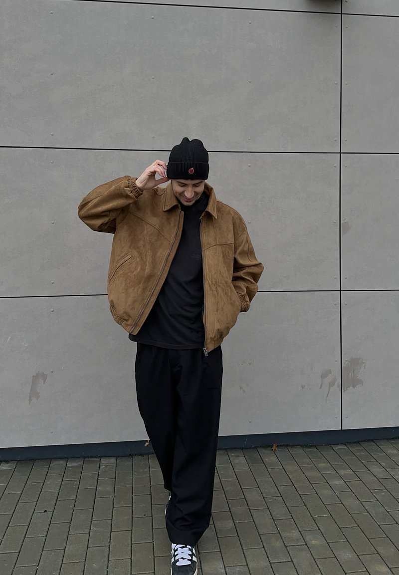 Veste en daim marron avec col, t-shirt noir, pantalon noir, bonnet noir et baskets grises, debout contre un mur gris.