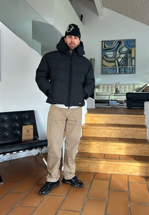 Hombre con chaqueta acolchada negra, gorro negro con estampado, pantalones beige y zapatos negros, de pie en el interior cerca de escaleras de madera y mobiliario moderno de sala.