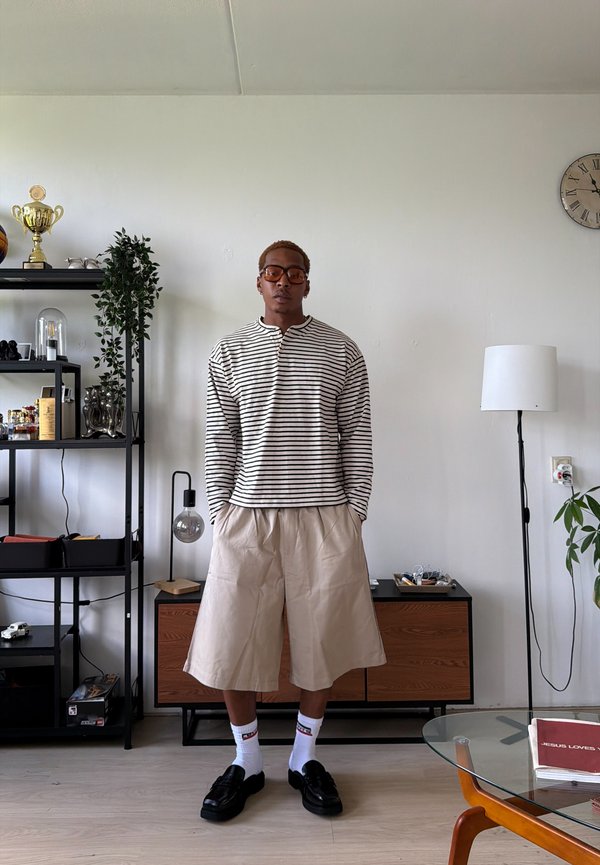 Junge Person mit Brille, gestreiftem Langarmshirt, beigen weiten Shorts, weißen Socken und schwarzen Loafers steht in einem modernen Wohnzimmer.