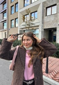 Manteau en fourrure marron, pull rose, écharpe blanche, lunettes de soleil rondes avec un motif écaille de tortue. Cheveux longs, tenant des lunettes de soleil. Arrière-plan urbain.