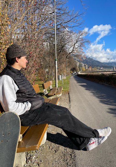 Un skater sentado en un banco de madera, vistiendo un chaleco acolchado negro, una camiseta de manga larga gris, pantalones negros y zapatillas blancas con acentos rojos.