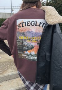 Smeđa majica s grafikom planinskog krajolika, s žutim i bijelim tekstom. Preko slojevito crna kožna jakna i karirana košulja ispod.